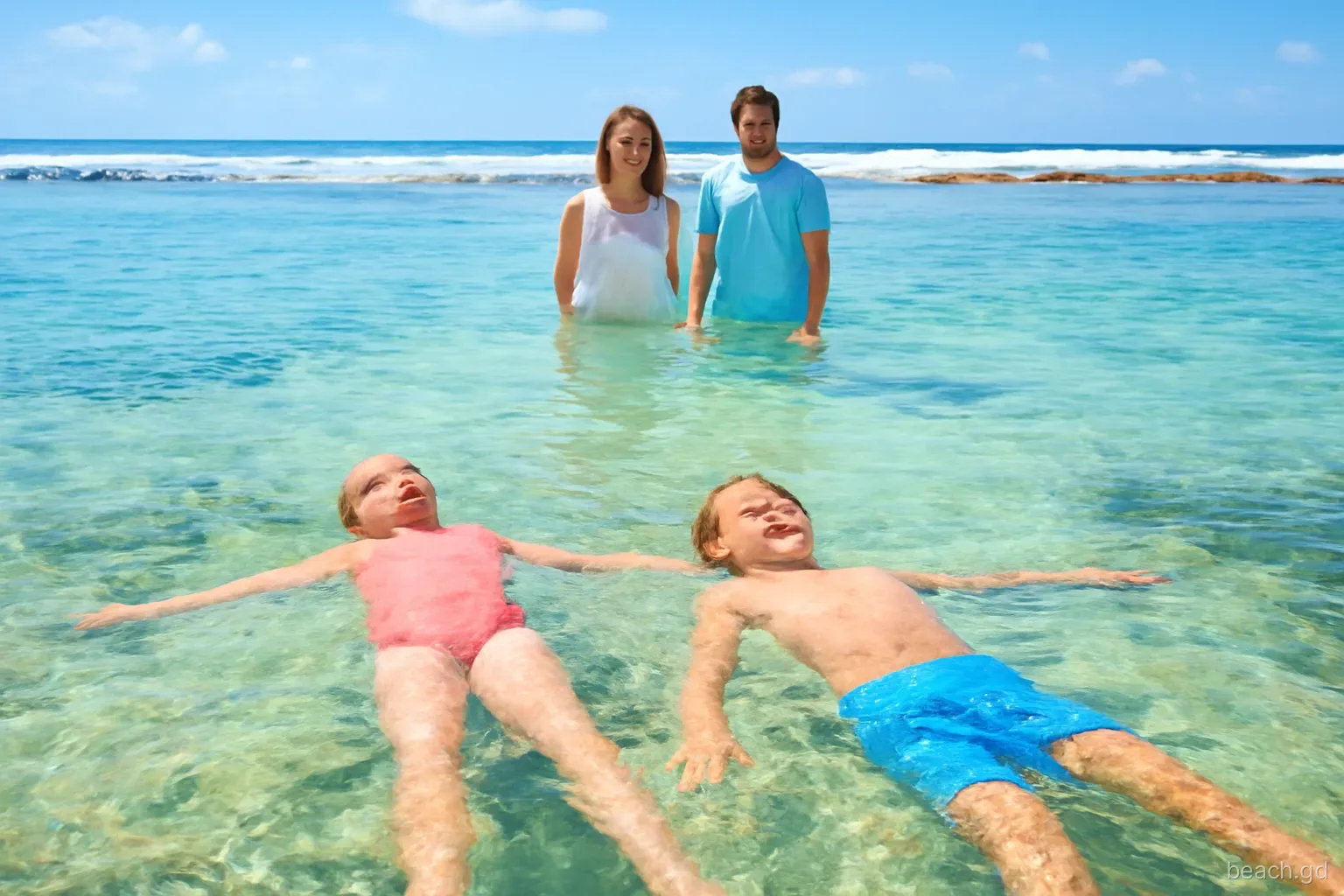Children floating in a shallow reef-sheltered lagoon at Bathway Beach, Grenada, white surf breaking on the outer reef behind, parents standing waist-deep, bright morning