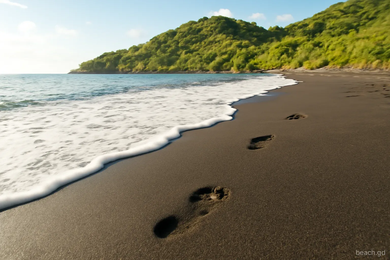 Close-up low angle of dark volcanic sand and foamy surf at a quiet cove on Grenada's leeward coast, green hillside behind, bare footprints, warm late afternoon light